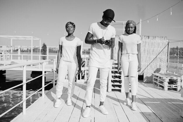 Tres elegantes amigos afroamericanos visten ropa blanca en el muelle de la playa Moda callejera de jóvenes negros Hombre negro con dos niñas africanas