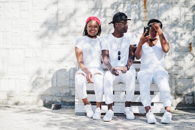 Tres elegantes amigos afroamericanos visten ropa blanca sentados en un banco Moda callejera de jóvenes negros Hombre negro con dos niñas africanas