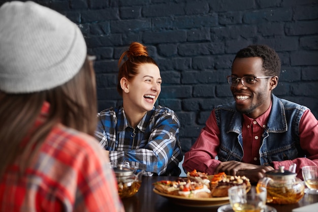 Tres elegantes jóvenes divirtiéndose durante el almuerzo en el café, hablando entre ellos y riéndose de chistes