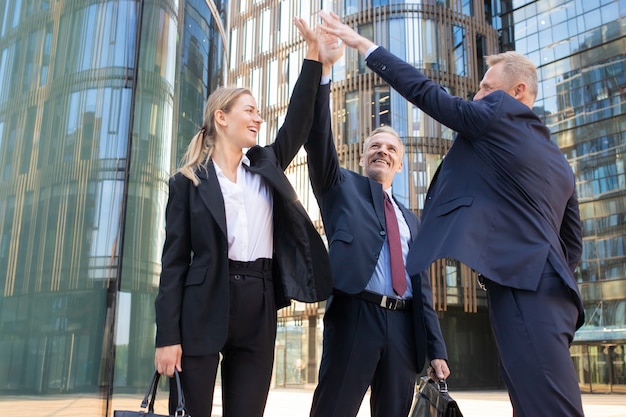 Foto gratuita tres empresarios alegres dando cinco y sonriendo. compañeros felices confiados celebrando un trato exitoso juntos, de pie al aire libre y levantando la mano. concepto de colaboración y trabajo en equipo