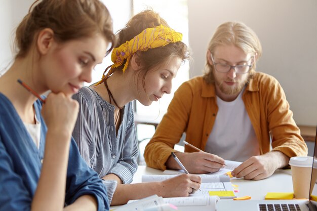 Tres estudiantes sentados juntos en su lugar de trabajo, escribiendo con lápices y estudiando literatura científica, preparándose para los exámenes en la universidad. Chico barbudo y dos mujeres trabajando en proyecto