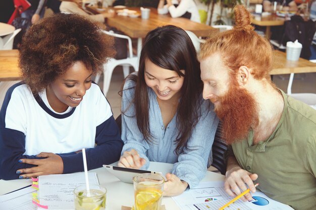 Tres estudiantes trabajando juntos en la tarea del hogar, sentados en la cafetería, investigando, navegando por Internet, usando wi-fi en el panel táctil.