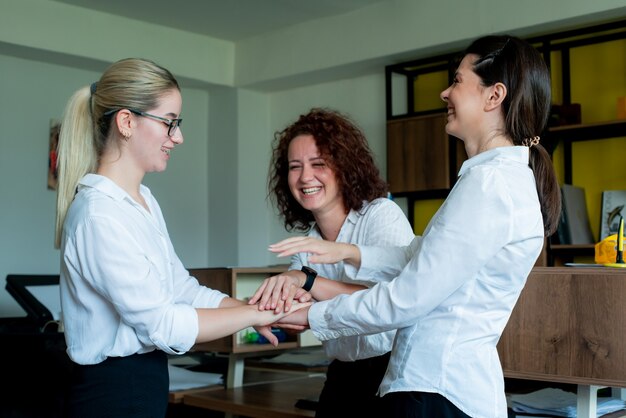 Tres felices compañeros de trabajo de oficina de mujeres satisfechas sonriendo alegremente apilaron sus manos juntas gesto de amistad, unidad y asociación en negocios de pie en la oficina