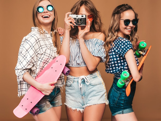 Tres hermosas chicas sonrientes con estilo con coloridas patinetas de centavo. Mujeres en ropa de camisa a cuadros de verano posando. Tomar fotos en la cámara de fotos retro