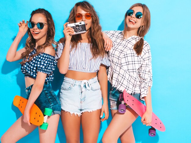 Tres hermosas chicas sonrientes con estilo con coloridas patinetas de centavo. Mujeres en ropa de camisa a cuadros de verano. Tomar fotos en la cámara de fotos retro