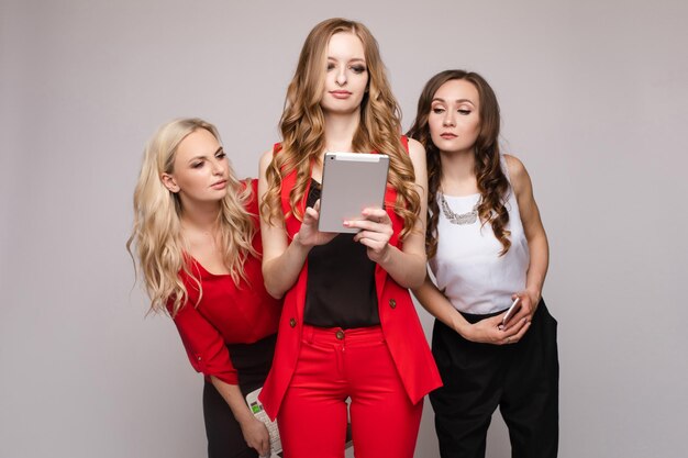 Tres hermosas y elegantes mujeres jóvenes vestidas de forma informal usando un teléfono móvil portátil y una tableta. Están usando aparatos para el trabajo y la comunicación.