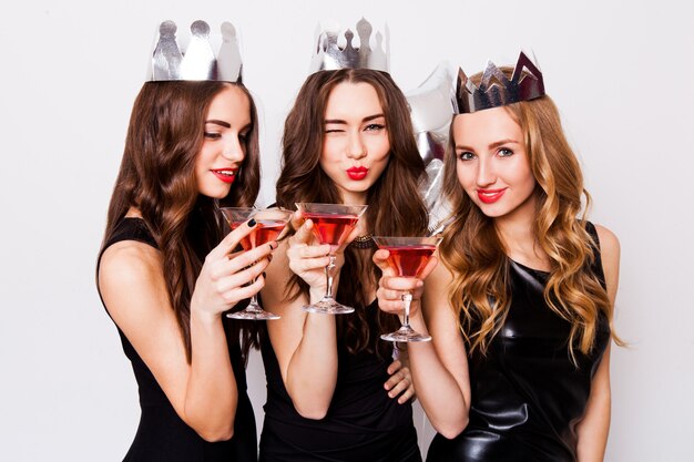 Tres hermosas mujeres elegantes celebran despedidas de soltera y beben cócteles. Las mejores amigas con vestido de noche negro, corona en la cabeza y anteojos. Maquillaje brillante, labios rojos. Dentro.