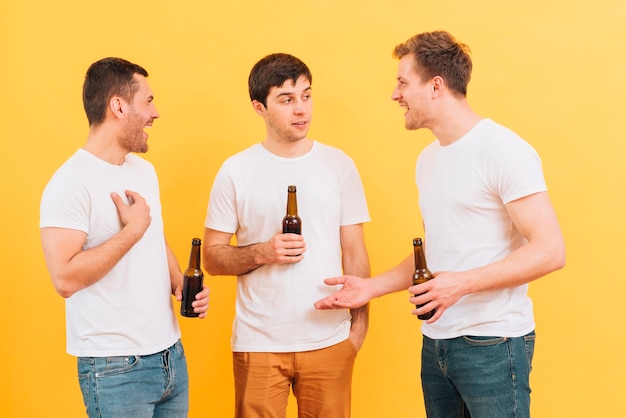 Tres jóvenes amigos varones disfrutando de la cerveza de pie contra el fondo amarillo