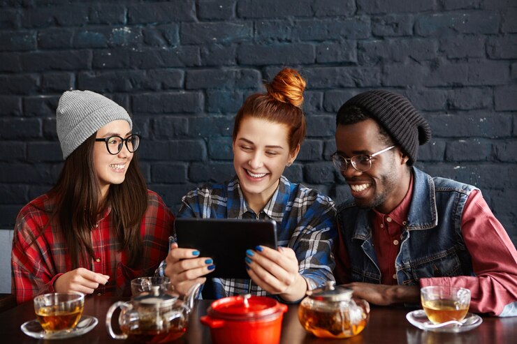 Foto gratuito tres jóvenes elegantes de diferentes razas viendo videos en línea en una tableta digital genérica mientras cenan juntos en el restaurante