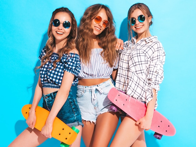 Tres jóvenes con estilo sonrientes hermosas chicas con patinetas coloridas centavo. Mujeres en ropa de verano posando en gafas de sol. Modelos positivos divirtiéndose