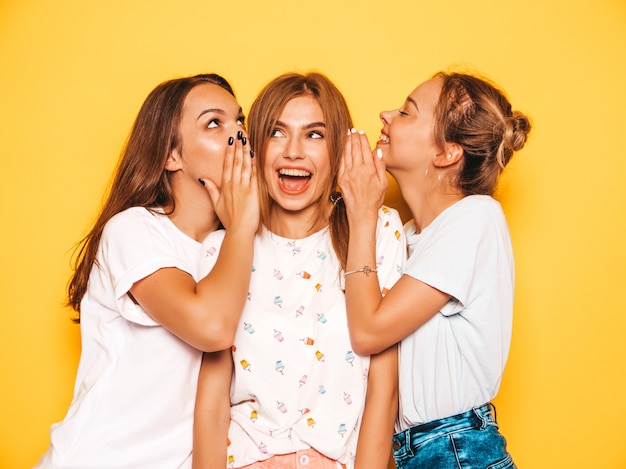 Tres jóvenes hermosas chicas hipster sonrientes en ropa de moda de verano. Mujeres despreocupadas atractivas que presentan cerca de la pared amarilla. Modelos positivos que se vuelven locos y se divierten. Compartan secretos, chismes