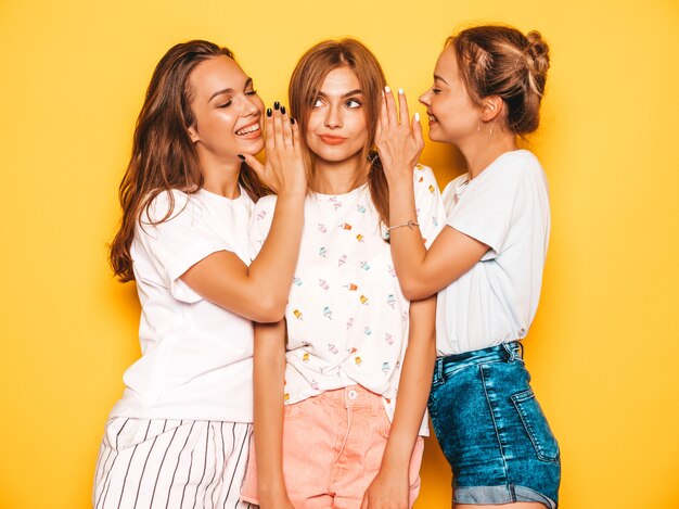 Tres jóvenes hermosas chicas hipster sonrientes en ropa de moda de verano. Mujeres despreocupadas atractivas que presentan cerca de la pared amarilla. Modelos positivos que se vuelven locos y se divierten. Compartan secretos, chismes