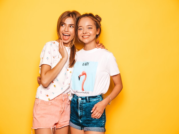 Tres jóvenes hermosas chicas hipster sonrientes en ropa de moda de verano. Mujeres despreocupadas atractivas que presentan cerca de la pared amarilla. Modelos positivos volviéndose locos y divirtiéndose. En gafas de sol