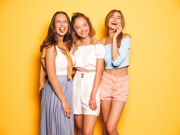 Tres jóvenes hermosas chicas hipster sonrientes en ropa de moda de verano. Mujeres despreocupadas atractivas que presentan cerca de la pared amarilla. Modelos positivos volviéndose locos y divirtiéndose