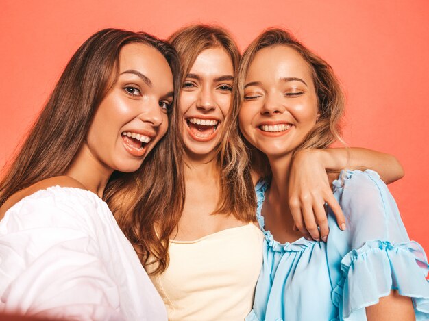Tres jóvenes hermosas chicas hipster sonrientes en ropa de moda de verano. Mujeres despreocupadas atractivas que presentan cerca de la pared rosada. Modelos positivos volviéndose locos. Tomar fotos de autorretrato autofoto en el teléfono inteligente