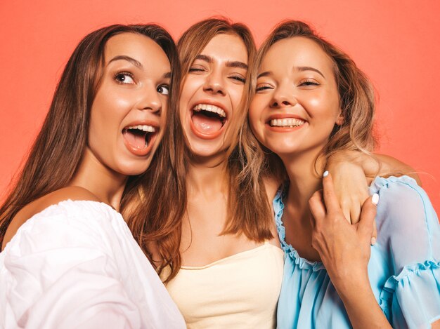 Tres jóvenes hermosas chicas hipster sonrientes en ropa de moda de verano. Mujeres despreocupadas atractivas que presentan cerca de la pared rosada. Modelos positivos volviéndose locos. Tomar fotos de autorretrato autofoto en el teléfono inteligente