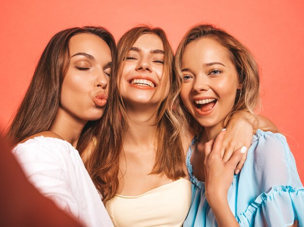 Tres jóvenes hermosas chicas hipster sonrientes en ropa de moda de verano. Mujeres despreocupadas atractivas que presentan cerca de la pared rosada. Modelos positivos volviéndose locos. Tomar fotos de autorretrato autofoto en el teléfono inteligente