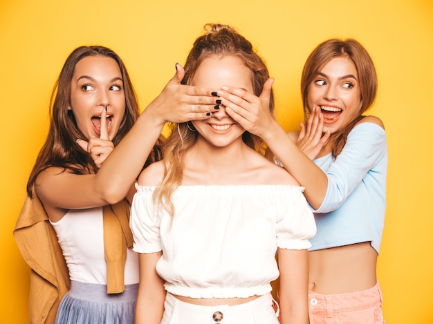 Tres jóvenes hermosas chicas hipster sonrientes en ropa de moda de verano. Mujeres despreocupadas y sexy posando junto a la pared amarilla. Modelos sorprendiendo a su amiga. Se cubren los ojos y se abrazan por detrás