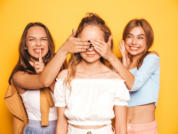 Tres jóvenes hermosas chicas hipster sonrientes en ropa de moda de verano. mujeres despreocupadas y sexy posando junto a la pared amarilla. modelos sorprendiendo a su amiga. se cubren los ojos y se abrazan por detrás