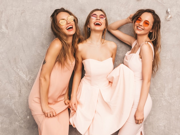 Tres jóvenes hermosas chicas sonrientes en moda rosa claro vestidos de verano. Sexy mujer despreocupada posando. Modelos positivos en gafas de sol redondas divirtiéndose