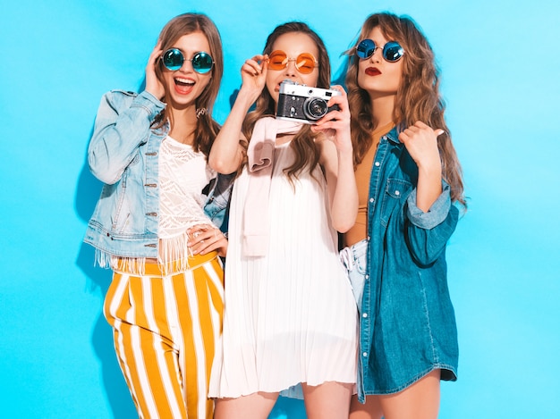 Tres jóvenes hermosas chicas sonrientes en moda verano coloridos vestidos y gafas de sol. Sexy mujer despreocupada posando. Tomar fotos en cámara retro