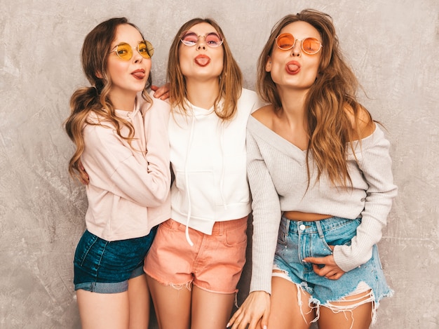 Tres jóvenes hermosas chicas sonrientes en ropa deportiva de moda de verano. Sexy mujer despreocupada posando. Modelos positivos en gafas de sol redondas divirtiéndose. Mostrando su lengua