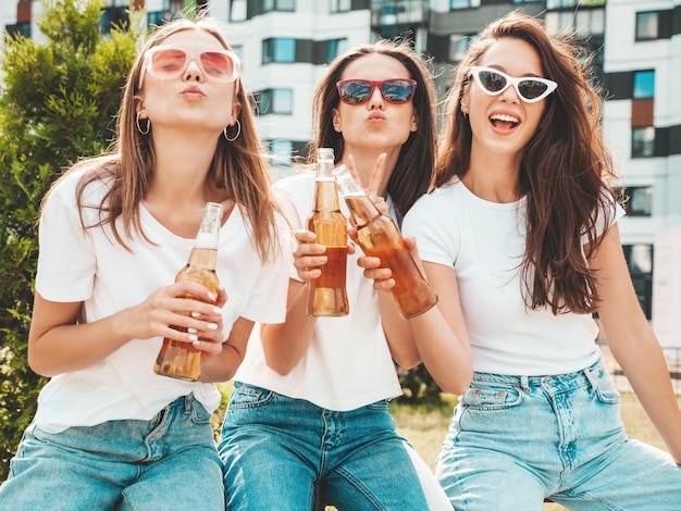 Tres jóvenes hermosas mujeres hipster sonrientes en la misma ropa de verano de moda Mujeres sexy despreocupadas posando en la calle Modelos positivos divirtiéndose con gafas de sol Bebiendo cerveza de botella Oktoberfest