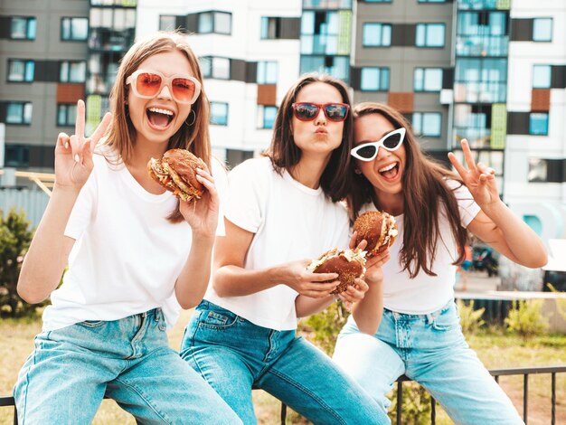 Tres jóvenes hermosas mujeres hipster sonrientes con la misma ropa de verano de moda Mujeres sexys y despreocupadas posando en la calle Modelos positivos divirtiéndose con gafas de sol Sosteniendo una hamburguesa jugosa y comiendo una hamburguesa