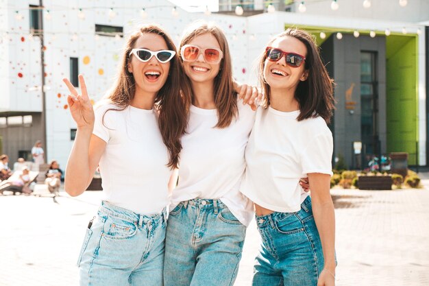 Tres jóvenes hermosas mujeres hipster sonrientes con la misma ropa de verano de moda Mujeres sexys y despreocupadas posando en el fondo de la calle Modelos positivos divirtiéndose con gafas de sol Abrazando Alegre y feliz