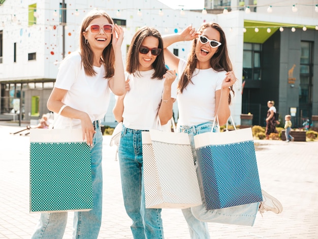 Tres jóvenes hermosas mujeres hipster sonrientes con la misma ropa de verano de moda Mujeres sexys y despreocupadas posando en el fondo de la calle Modelos positivos divirtiéndose con gafas de sol Abrazando Sosteniendo bolsas de compras