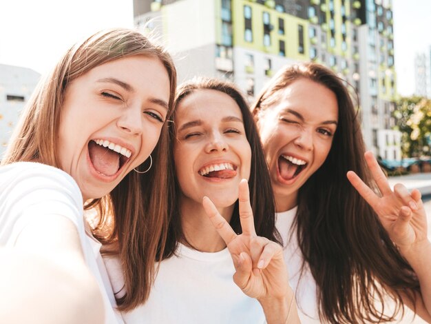 Tres jóvenes hermosas mujeres hipster sonrientes con la misma ropa de verano de moda Mujeres sexys y despreocupadas posando en el fondo de la calle Modelos positivos divirtiéndose con gafas de sol Tomando fotos autofotos de Pov
