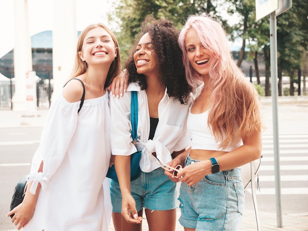 Tres jóvenes hermosas mujeres hipster sonrientes con ropa de verano de moda. Mujeres multirraciales sexy y despreocupadas posando en el fondo de la calle. Modelos positivos divirtiéndose con gafas de sol. Alegres y felices.