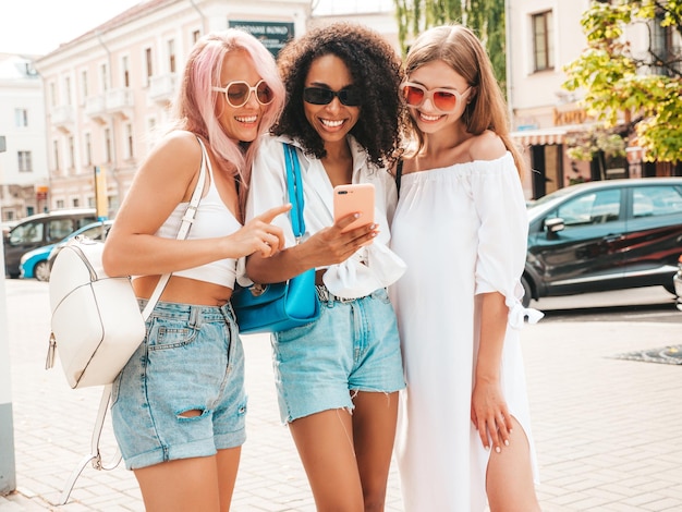 Tres jóvenes hermosas mujeres hipster sonrientes con ropa de verano de moda. Mujeres multirraciales sexys y despreocupadas posando en la calle. Modelos positivos con gafas de sol Mirando la pantalla del teléfono inteligente Usando aplicaciones telefónicas