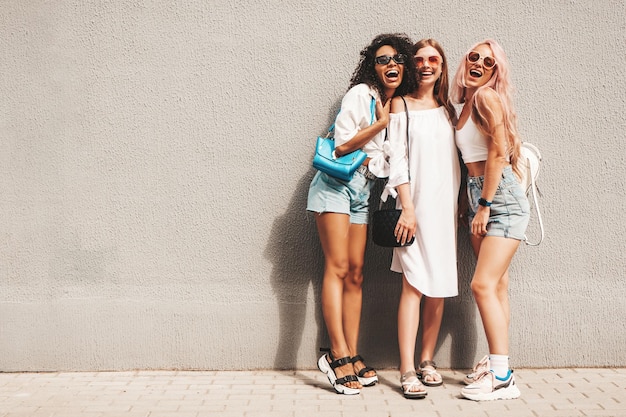 Tres jóvenes hermosas mujeres sonrientes en ropa de verano de moda Mujeres multirraciales despreocupadas sexy posando en el fondo de la calle cerca de la pared Modelos positivos divirtiéndose con gafas de sol Alegre y feliz