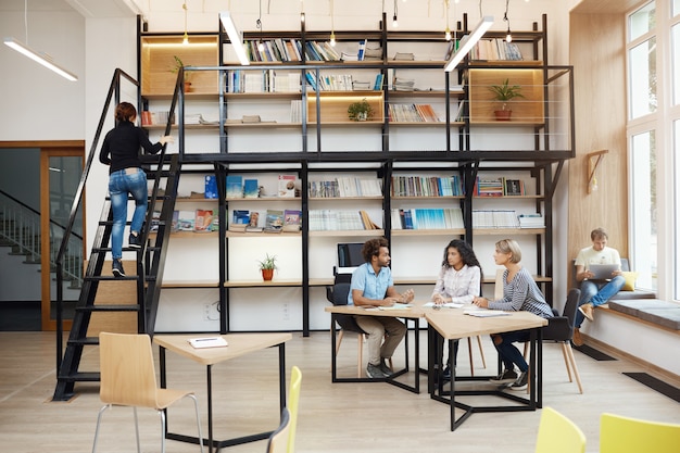 Tres jóvenes principiantes en perspectiva sentados en una moderna biblioteca de luz en una reunión, hablando sobre un nuevo proyecto, buscando detalles del trabajo, teniendo un día productivo en un ambiente de amigos,