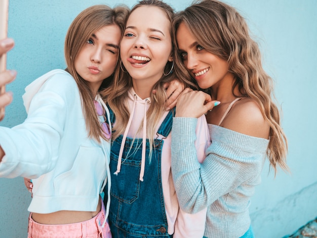 Tres jóvenes sonrientes mujeres hipster en ropa de verano. Chicas tomando fotos de autorretrato en smartphone. Modelos posando en la calle cerca de la pared. Mujer mostrando emociones positivas. Muestra lengua.