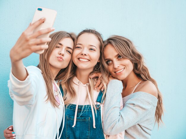 Tres jóvenes sonrientes mujeres hipster en ropa de verano. Chicas tomando fotos de autorretrato en smartphone. Modelos posando en la calle cerca de la pared. Mujeres mostrando emociones positivas.