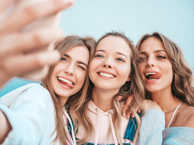 Tres jóvenes sonrientes mujeres hipster en ropa de verano. Chicas tomando fotos de autorretrato en smartphone. Modelos posando en la calle cerca de la pared. Mujeres mostrando emociones positivas.