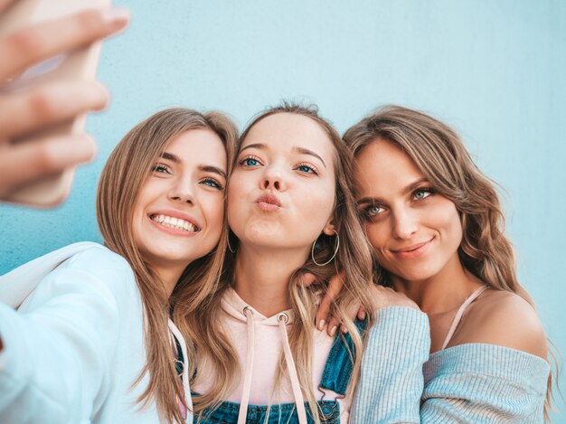 Tres jóvenes sonrientes mujeres hipster en ropa de verano. Chicas tomando fotos de autorretrato en smartphone. Modelos posando en la calle cerca de la pared. Mujeres mostrando emociones positivas.