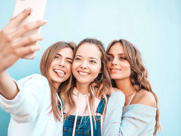 Tres jóvenes sonrientes mujeres hipster en ropa de verano. Chicas tomando fotos de autorretrato en smartphone. Modelos posando en la calle cerca de la pared. Mujeres mostrando emociones positivas.