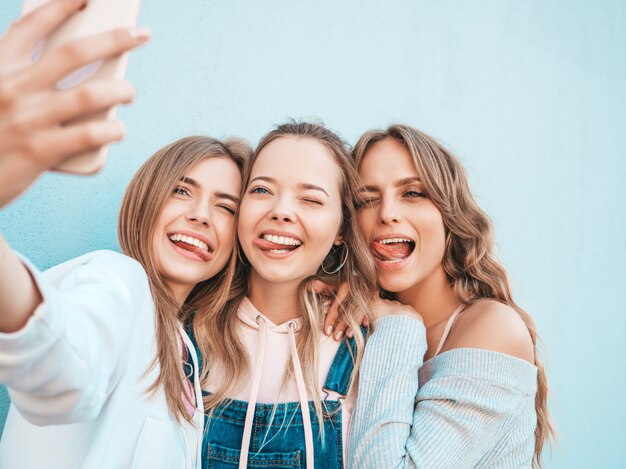 Tres jóvenes sonrientes mujeres hipster en ropa de verano. Chicas tomando fotos de autorretrato en smartphone. Modelos posando en la calle cerca de la pared. Mujeres mostrando emociones positivas.