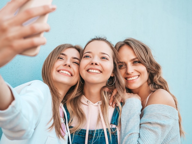 Tres jóvenes sonrientes mujeres hipster en ropa de verano. Chicas tomando fotos de autorretrato en smartphone. Modelos posando en la calle cerca de la pared. Mujeres mostrando emociones positivas.