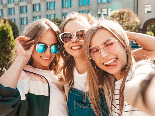 Tres jóvenes sonrientes mujeres hipster en ropa de verano. Chicas tomando fotos de autorretrato en smartphone. Modelos posando en la calle. Mujeres mostrando emociones positivas en la cara.