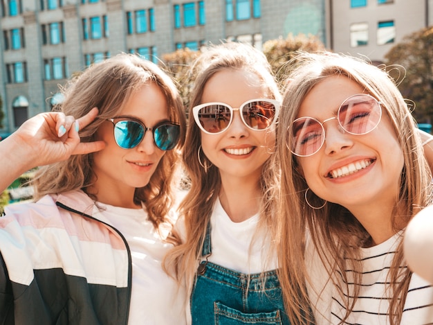 Tres jóvenes sonrientes mujeres hipster en ropa de verano. Chicas tomando fotos de autorretrato en smartphone. Modelos posando en la calle. Mujeres mostrando emociones positivas en la cara.