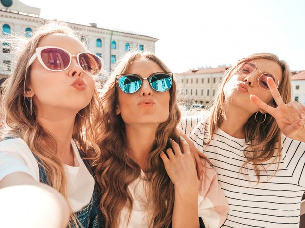Tres jóvenes sonrientes mujeres hipster en ropa de verano. Chicas tomando fotos de autorretrato en smartphone. Modelos posando en la calle. Mujeres mostrando emociones positivas en la cara.