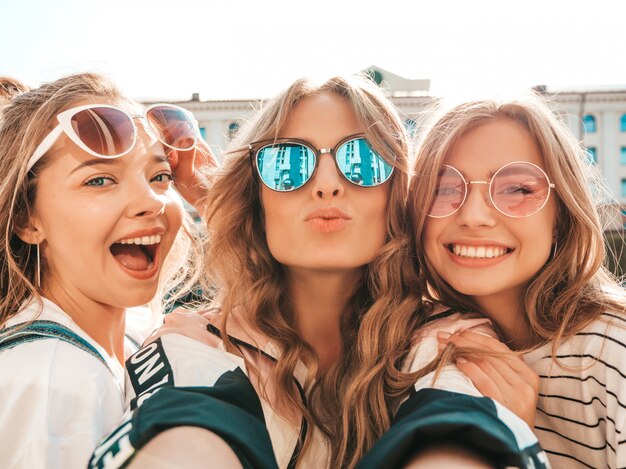 Tres jóvenes sonrientes mujeres hipster en ropa de verano. Chicas tomando fotos de autorretrato en smartphone. Modelos posando en la calle. Mujeres mostrando emociones positivas en las gafas de sol.