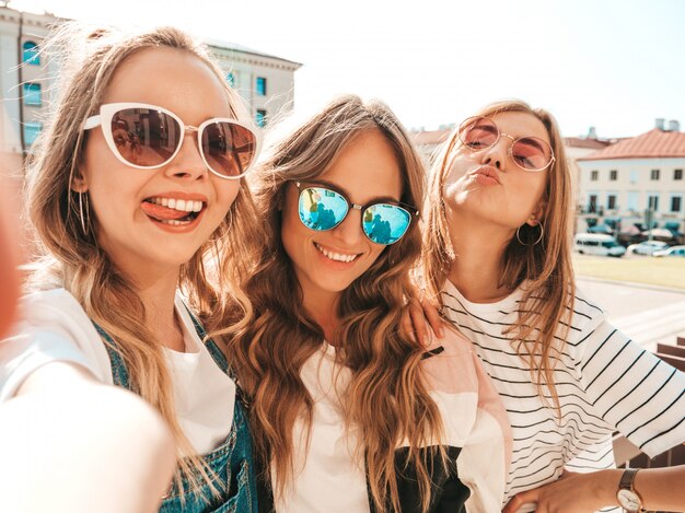 Tres jóvenes sonrientes mujeres hipster en ropa de verano. Chicas tomando fotos de autorretrato en smartphone. Modelos posando en la calle. Mujeres mostrando emociones positivas en las gafas de sol.