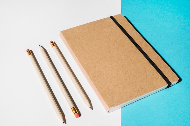 Tres lápices y cuaderno cerrado con tapa marrón.