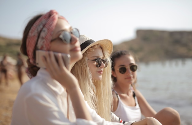 Tres mujeres jóvenes con gafas en la playa, una hablando por teléfono