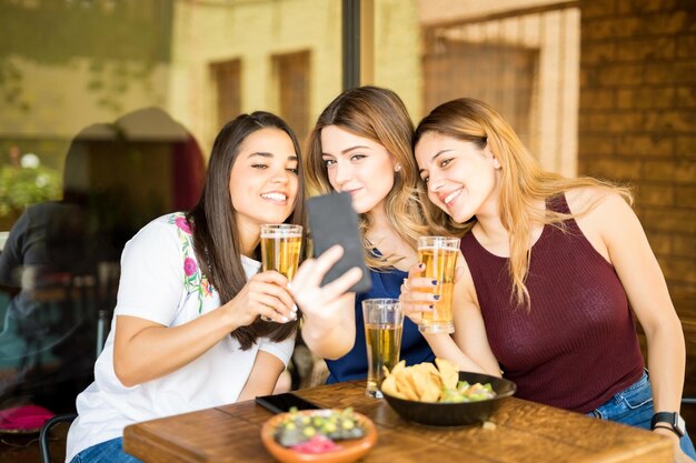 Tres mujeres jóvenes sentadas en el restaurante con cervezas tomando selfie con teléfono móvil